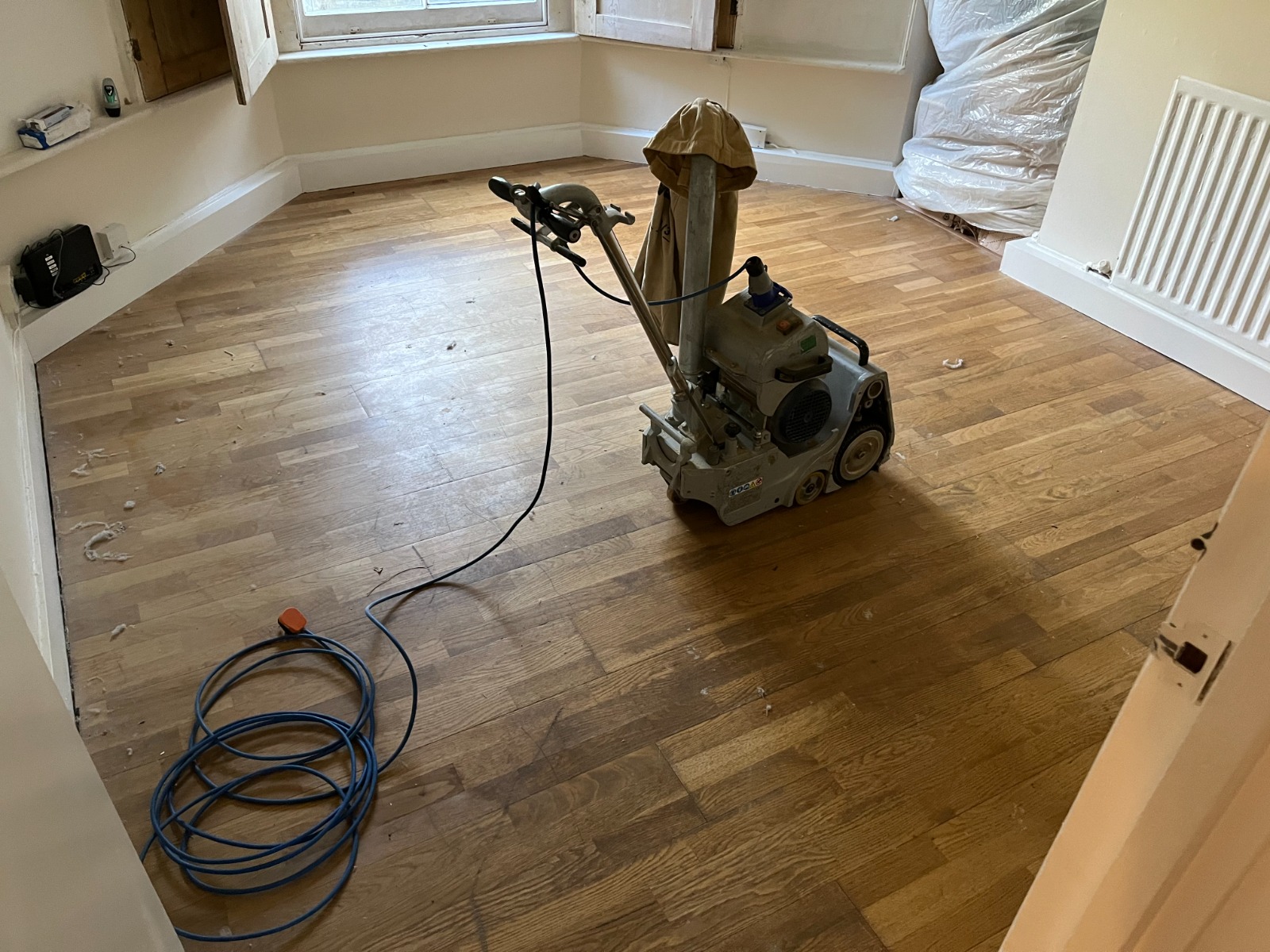 Professional drum sander with dust bag in use to Sand Floors on an oak strip hardwood floor, mid-preparation for refinishing inside a cleared room.