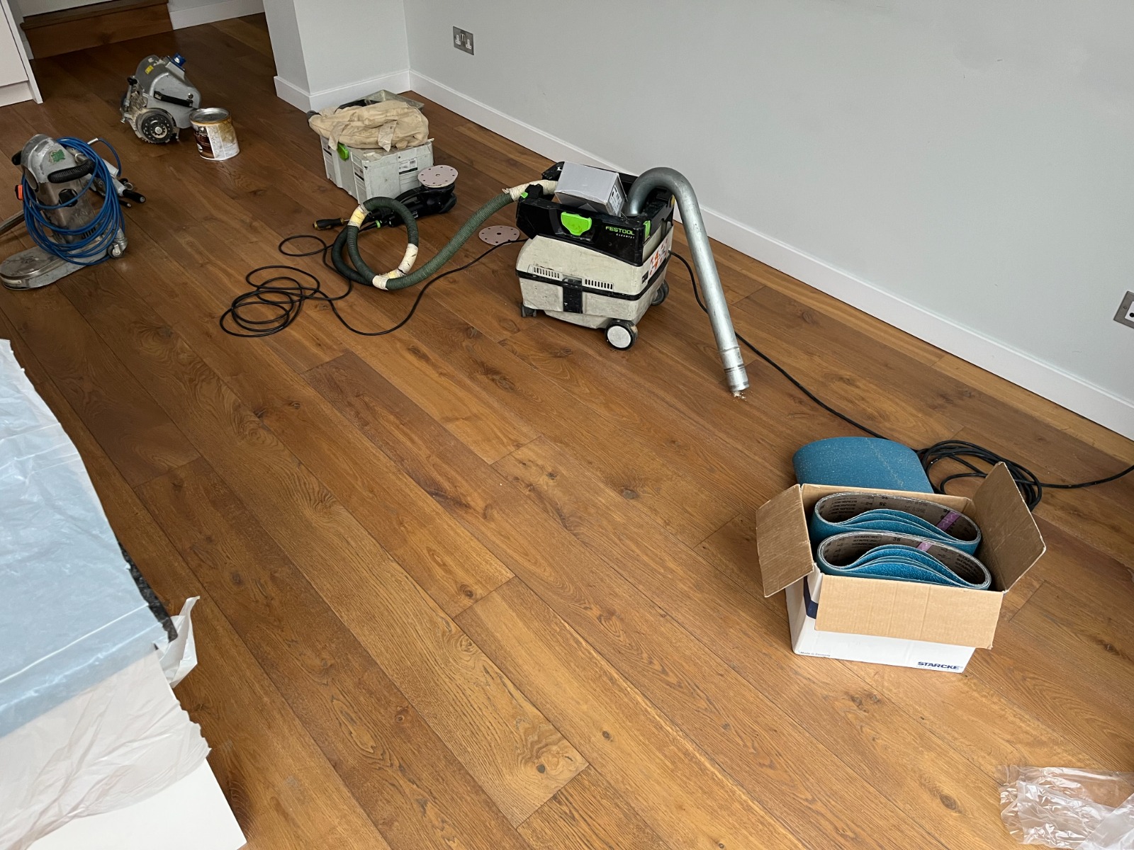 Professional setup to Sand Floors on oak wide-plank engineered hardwood, with edger, buffer, HEPA dust-extraction unit and boxes of sanding belts laid out.